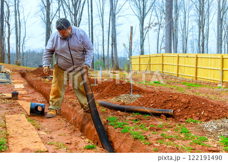 Worker works diligently to install drainage system in his backyard, surrounded by trees fence for enhance water flow management 122191490
