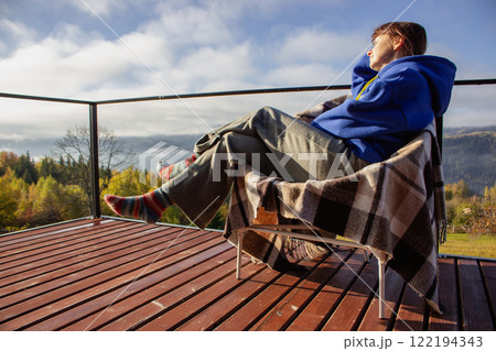 Woman sitting on terrace with checkered plaid and knitted socks on mountains background. Tranquil relax in mountains. Happy girl sitting on wooden terrace. Vacations in Ukraine. Chill in Carpathians. Woman sitting on terrace with checkered plaid and knitted socks on mountains background. Tranquil relax in mountains. Happy girl sitting on wooden terrace. Vacations in Ukraine. Chill in Carpathians. 122194343