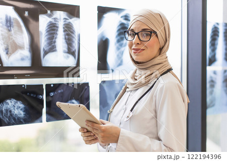 Confident Muslim woman doctor examines x-rays in hospital. She is in her 30s, wearing a hijab and glasses, holding a tablet. Image conveys professionalism and healthcare expertise. Confident Muslim woman doctor examines x-rays in hospital. She is in her 30s, wearing a hijab and glasses, holding a tablet. Image conveys professionalism and healthcare expertise. 122194396