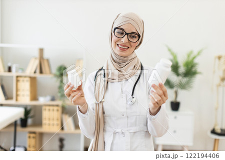 Confident Muslim female doctor holding medication bottle and blister in bright clinic. Young adult woman with glasses. Medical professional promotes health and treatment. Caucasian race. Confident Muslim female doctor holding medication bottle and blister in bright clinic. Young adult woman with glasses. Medical professional promotes health and treatment. Caucasian race. 122194406