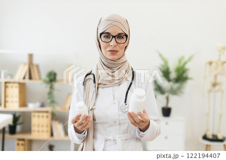 Muslim female doctor in her 30s holding white medication bottles in hands. She is wearing a hijab and lab coat, conveying professionalism and healthcare concept in bright modern office. Muslim female doctor in her 30s holding white medication bottles in hands. She is wearing a hijab and lab coat, conveying professionalism and healthcare concept in bright modern office. 122194407