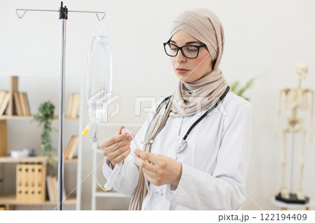 Young Muslim woman doctor sets up IV dropper in clinic. Focused healthcare professional, wearing hijab and stethoscope, demonstrates meticulous medical preparation. Young Muslim woman doctor sets up IV dropper in clinic. Focused healthcare professional, wearing hijab and stethoscope, demonstrates meticulous medical preparation. 122194409