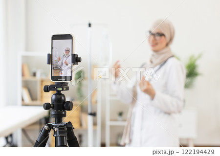 Young Muslim woman in medical attire records video tutorial about intravenous dropper setup using smartphone. Educational content creation in a healthcare environment with modern technology. Young Muslim woman in medical attire records video tutorial about intravenous dropper setup using smartphone. Educational content creation in a healthcare environment with modern technology. 122194429