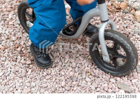 A young child riding a balance bike on a gravel pathway, wearing sturdy black shoes and blue pants. An active and playful moment, emphasizing outdoor activity, childhood exploration, and learning. A young child riding a balance bike on a gravel pathway, wearing sturdy black shoes and blue pants. An active and playful moment, emphasizing outdoor activity, childhood exploration, and learning. 122195398