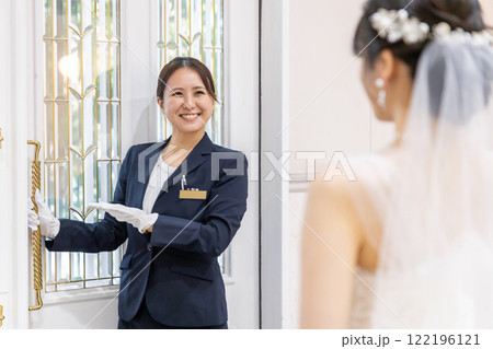 結婚式場のドアを開けるウェディングプランナー・ドアスタッフ・ホテリエの女性と新郎新婦（結婚式） 122196121
