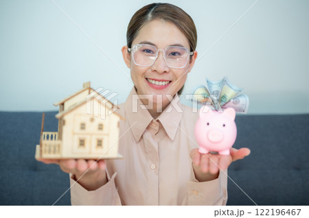 Asian woman holding a small mock up house with piggy bank on her hands. Saving money can help you reach your goals. Asian woman holding a small mock up house with piggy bank on her hands. Saving money can help you reach your goals. 122196467