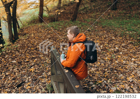 Blonde young man with thermos of tea wearing backpack standing on autumn forest trail, hiking alone. Freedom and active lifestyle concept. High quality photo 122198532