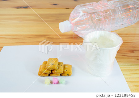 ビニール袋でカバーされた水が入った紙コップと避難所配給品の一食分の乾パン ビニール袋でカバーされた水が入った紙コップと避難所配給品の一食分の乾パン 122199458