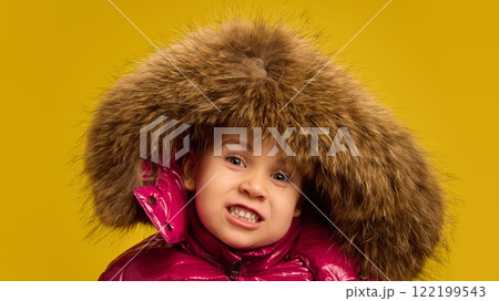 Child, girl in shiny pink winter coat with fur hood, smiling, making funny playful face against yellow studio background. Child, girl in shiny pink winter coat with fur hood, smiling, making funny playful face against yellow studio background. 122199543