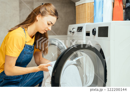 Tired woman sitting washing machine inside home laundry on bathroom floor and looking at dirty bed linen. Angry female fatigue, depressed and crisis for cleaning challenge, disaster and problem 122200144