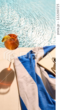 Vibrant orange drink with lemon slice near shimmering pool. Blue and white towel with sunglasses and open book suggest calm holiday moments. Vibrant orange drink with lemon slice near shimmering pool. Blue and white towel with sunglasses and open book suggest calm holiday moments. 122200725