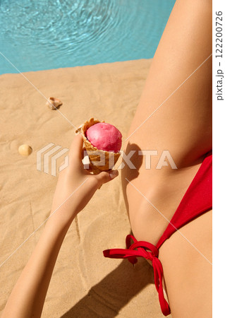 Hand holding pink ice cream cone, red bikini, and sandy background near pool, capturing sweet and carefree summer holiday moments by sparkling water. Hand holding pink ice cream cone, red bikini, and sandy background near pool, capturing sweet and carefree summer holiday moments by sparkling water. 122200726