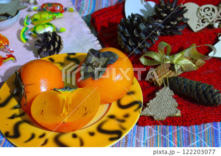 Still life of ripe bright persimmons, pine cones and candies on a crimson woolen background. Fruits lie on a beautiful plate. Whole and cut fruits. 122203077