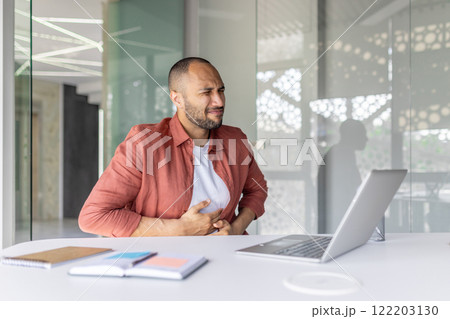 A professional seated at a desk appears in discomfort while holding his stomach, expressing pain. The modern office environment emphasizes his professional context and the seriousness of situation. 122203130