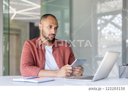 A man holds a tablet while working at a desk with a laptop and notebook in a contemporary office space. He appears focused and immersed in his task. A man holds a tablet while working at a desk with a laptop and notebook in a contemporary office space. He appears focused and immersed in his task. 122203138