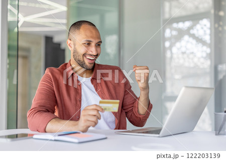 A cheerful man celebrates a successful online transaction by utilizing a credit card and laptop in a bright office setting. The image conveys concepts of digital technology, satisfaction. 122203139