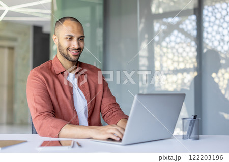 A professional man using a laptop while seated at a desk in a bright, modern workspace, exuding positivity and focus. The scene highlights productivity, business, and a pleasant working atmosphere. A professional man using a laptop while seated at a desk in a bright, modern workspace, exuding positivity and focus. The scene highlights productivity, business, and a pleasant working atmosphere. 122203196