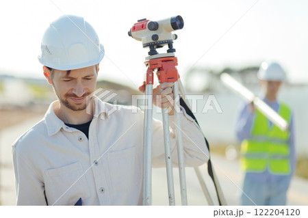 Engineer Surveyor Carries Tripod Level on shoulder. Walks at Construction Place Engineer Surveyor Carries Tripod Level on shoulder. Walks at Construction Place 122204120