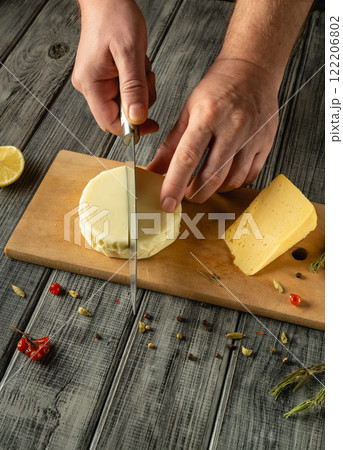 Hands expertly slice a round of cheese on a weathered wooden cutting board. Nearby, vibrant herbs, spices, and a lemon add color and flavor to the rustic kitchen atmosphere Hands expertly slice a round of cheese on a weathered wooden cutting board. Nearby, vibrant herbs, spices, and a lemon add color and flavor to the rustic kitchen atmosphere 122206802