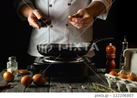 A skilled cook prepares a delicious meal by adding spices to a sizzling pan. Fresh eggs and ingredients adorn the rustic kitchen counter, creating an inviting atmosphere for culinary exploration 122206803
