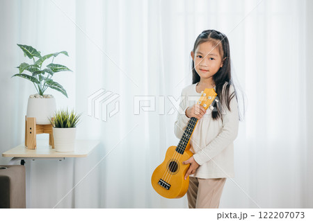 Asian girl strums a ukulele while standing in a cozy, well-decorated living room. Her cheerful expression and love for music harmonize perfectly with the modern, softly lit environment. 122207073