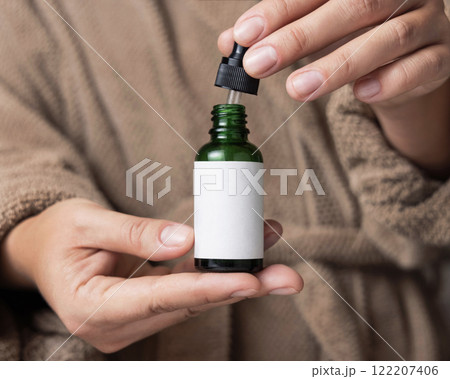 Hands in brown bathrobe opening green dropper bottle with label closeup, cosmetic mockup Hands in brown bathrobe opening green dropper bottle with label closeup, cosmetic mockup 122207406