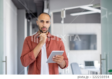 Portrait of a serious confident man with a tablet computer in his hands, businessman looking at the camera, standing inside the office at his workplace in casual clothes. Portrait of a serious confident man with a tablet computer in his hands, businessman looking at the camera, standing inside the office at his workplace in casual clothes. 122210354