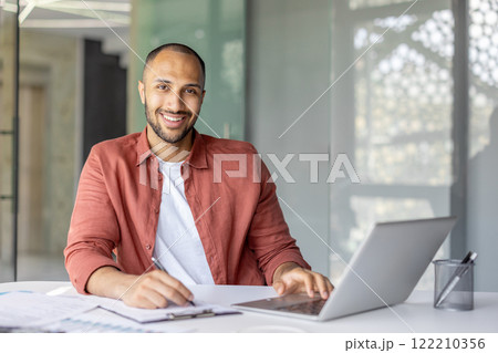 Portrait of a young successful businessman financier at his workplace inside the office, the employee is smiling and looking at the camera, writing down data on a paper report document Portrait of a young successful businessman financier at his workplace inside the office, the employee is smiling and looking at the camera, writing down data on a paper report document 122210356