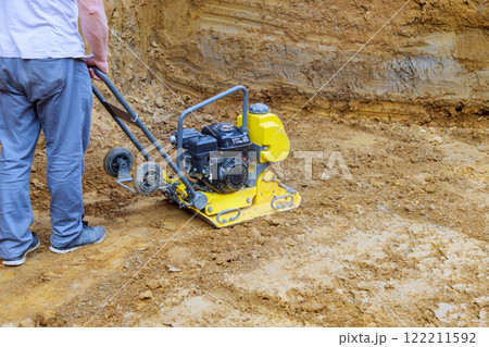 Contractor operates plate compactor on construction site, compacting soil for solid foundation. Contractor operates plate compactor on construction site, compacting soil for solid foundation. 122211592