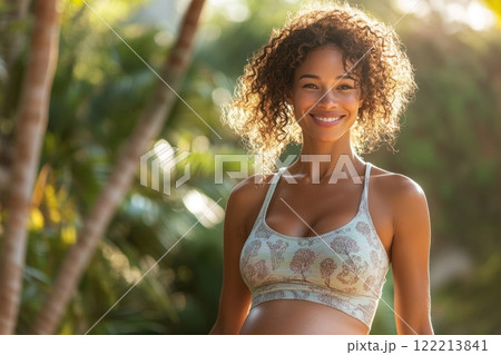 Pregnant woman with curly hair smiling warmly outdoors, wearing a patterned sports bra, surrounded by lush greenery and glowing in natural sunlight 122213841