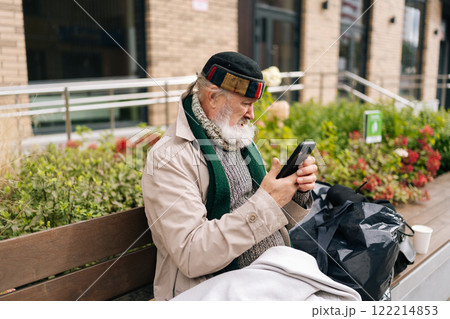 Side view of aged beggar man wrapped in blanket using smartphone sitting on urban bench outside building on cold autumn day. Concept of poverty and vulnerability, evoking empathy and compassion. 122214853
