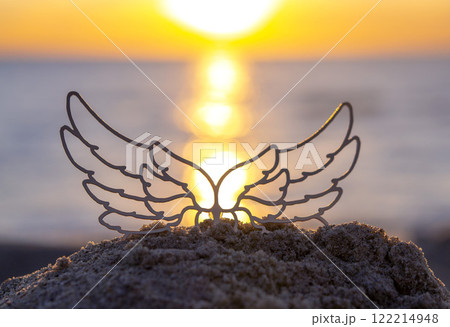 Black silhouette stick with transparent wings in sand on sandy beach of sea shore against sea background, sea waves and sun path during sunrise or sunset with rising or setting sun close up. Concept Black silhouette stick with transparent wings in sand on sandy beach of sea shore against sea background, sea waves and sun path during sunrise or sunset with rising or setting sun close up. Concept 122214948