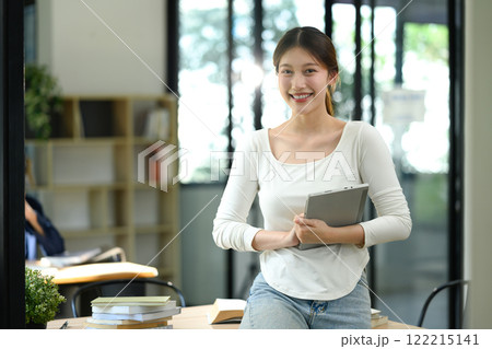 Portrait of cheerful university student holding digital tablet standing confidently in a modern study space Portrait of cheerful university student holding digital tablet standing confidently in a modern study space 122215141
