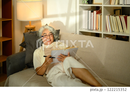 Relaxed an elderly woman in glasses lounging on a comfortable sofa and writing in a notebook Relaxed an elderly woman in glasses lounging on a comfortable sofa and writing in a notebook 122215352