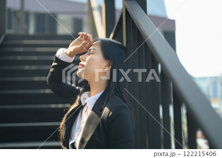 Stressed and exhausted businesswoman leaning against railing with hand on her forehead and closing her eyes Stressed and exhausted businesswoman leaning against railing with hand on her forehead and closing her eyes 122215406