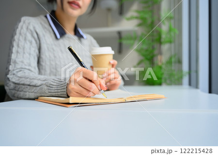 Close up of young woman writing in notebook while sitting in a bright, natural light filled space Close up of young woman writing in notebook while sitting in a bright, natural light filled space 122215428
