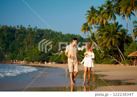 Romantic couple walks on beach, barefoot, holding coconut drinks, enjoying sunset. Man, woman in casual summer clothes, romantic stroll, tropical seaside, date concept. Happy pair, love, vacation. Romantic couple walks on beach, barefoot, holding coconut drinks, enjoying sunset. Man, woman in casual summer clothes, romantic stroll, tropical seaside, date concept. Happy pair, love, vacation. 122216347