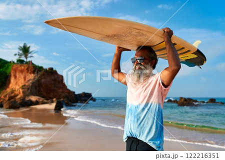 Senior man carries surfboard at beach. Active lifestyle for elderly. Grey-haired male enjoys ocean sport. Diverse mature surfer, vibrant life. Positive outlook, healthy aging. Senior man carries surfboard at beach. Active lifestyle for elderly. Grey-haired male enjoys ocean sport. Diverse mature surfer, vibrant life. Positive outlook, healthy aging. 122216351