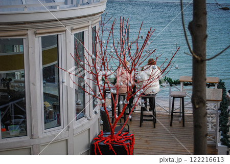 A plot shot of a seascape. A decorative red tree with small cups, behind which you can see people sitting, looking at the sea. A plot shot of a seascape. A decorative red tree with small cups, behind which you can see people sitting, looking at the sea. 122216613