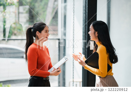Two women in vibrant sweaters chatting and smiling in a bright office environment, holding documents and enjoying a casual conversation. Two women in vibrant sweaters chatting and smiling in a bright office environment, holding documents and enjoying a casual conversation. 122220912