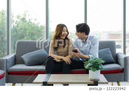 Young asian couple sitting on a couch, laughing together and sharing a mobile phone, in a bright living room. 122223942