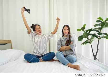 Two girl sit on a bed in a bright room. One girl raise her arm in excitement after winning a game, while the other appear annoyed, showcasing different reaction to gaming. Two girl sit on a bed in a bright room. One girl raise her arm in excitement after winning a game, while the other appear annoyed, showcasing different reaction to gaming. 122223973