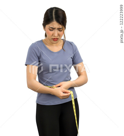 A woman stand with a tape measure around her waist, displaying a worried expression while assessing her body size. She wear a casual blue top and black pant. Portrait on white background. Isolated 122223999