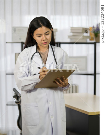 A medical professional in a white coat is writing note on a clipboard while standing in a clinic environment. This scene capture the dedication to patient care and documentation. 122224091