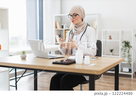 Muslim female doctor wearing hijab conducts video consultation with patient using laptop. Young adult in professional office setting discussing healthcare remotely, engaging in telemedicine. Muslim female doctor wearing hijab conducts video consultation with patient using laptop. Young adult in professional office setting discussing healthcare remotely, engaging in telemedicine. 122224480