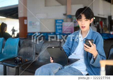 Young traveler sitting in a busy transport station and using smartphone during downtime. Young traveler sitting in a busy transport station and using smartphone during downtime. 122227708