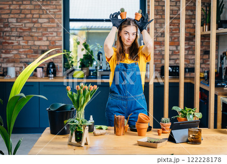 Having Fun Growing Plants Concept. Playful young female holds small succulents plants in pots like ears at cozy home with modern kitchen interior on background. 122228178