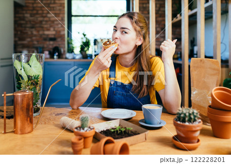 Searching for best advice. Romantic brunette haired smiling female gardener eats sweet cake, drinking coffee sitting surround her plants pots, equipment, tools, watering can in home garden at home. Searching for best advice. Romantic brunette haired smiling female gardener eats sweet cake, drinking coffee sitting surround her plants pots, equipment, tools, watering can in home garden at home. 122228201