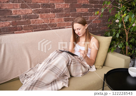 Young caucasian adult female relaxing on sofa with tablet and blanket in cozy 122228543