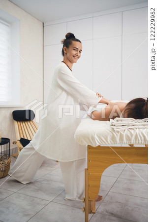 Massage process. Beautiful smiling woman in white outfit makes body massage to lying client in pro salon, vertical image, selective focus. 122229982